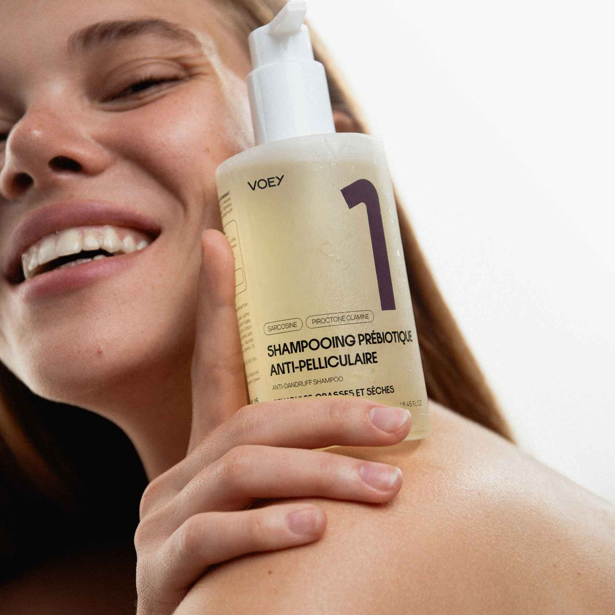 A smiling woman holds a bottle of VoeY shampoo against her shoulder. The bottle is frosted and filled with a pale yellow liquid, featuring a white pump and a large purple number 1 on the label.