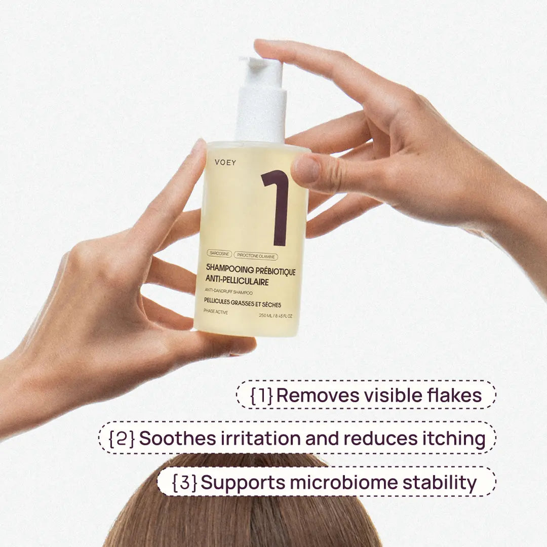 A person holds a bottle of VOEY Step 1 shampoo against a white background, with text overlays describing benefits: removes visible flakes, soothes irritation and reduces itching, and supports microbiome stability.