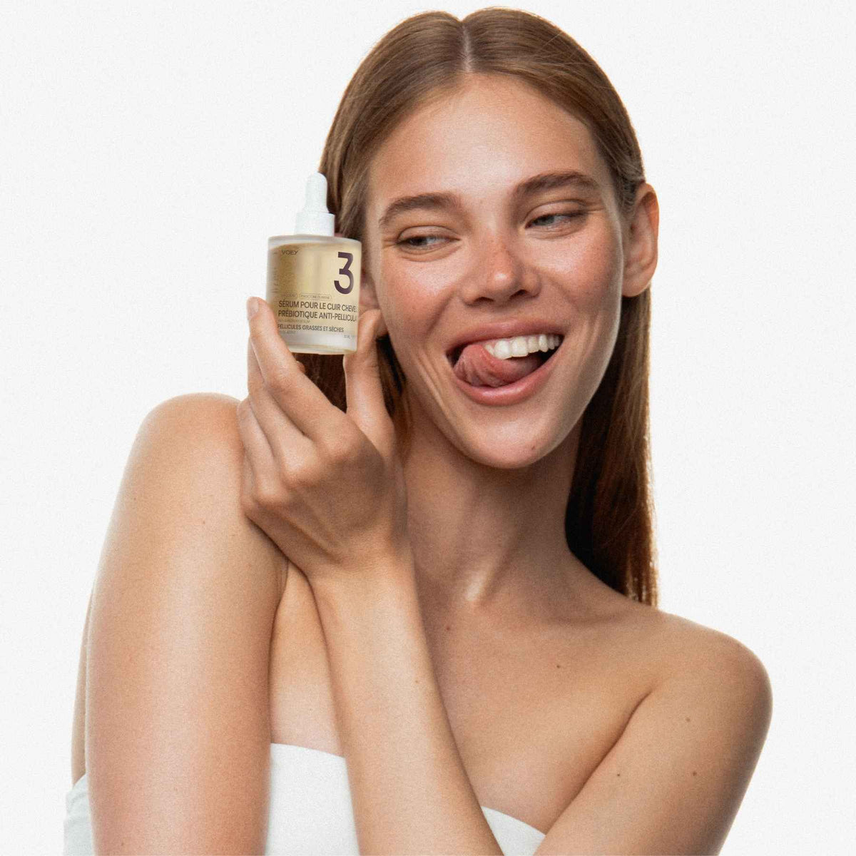 A smiling woman with long light brown hair holds a small cosmetic jar near her face against a clean white background. The jar appears to be a gold-toned skincare bottle with a white dropper cap and a visible number 3 label.