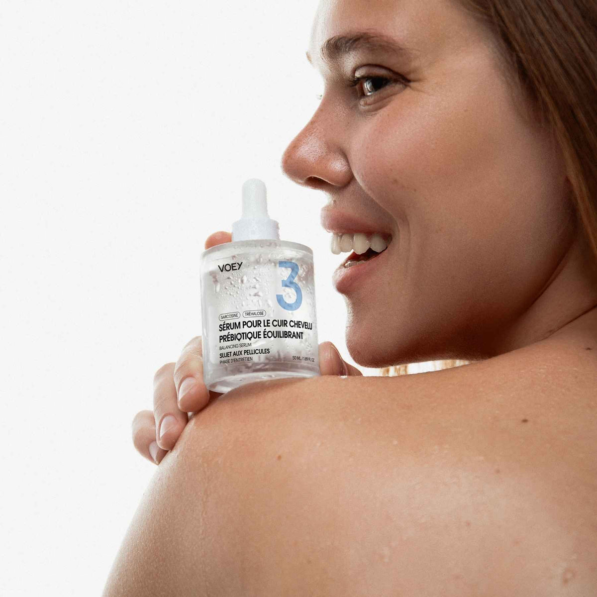 A close-up lifestyle shot of a woman holding a clear bottle of VOOEY 3-Step Skin Rescue serum on her shoulder against a bright white background.