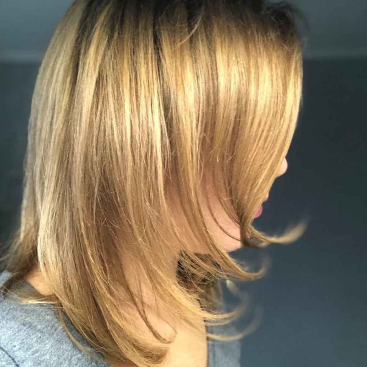 Side profile of a person with shoulder-length layered blonde hair, showing warm golden and light brown highlights, wearing a gray top against a dark gray background.