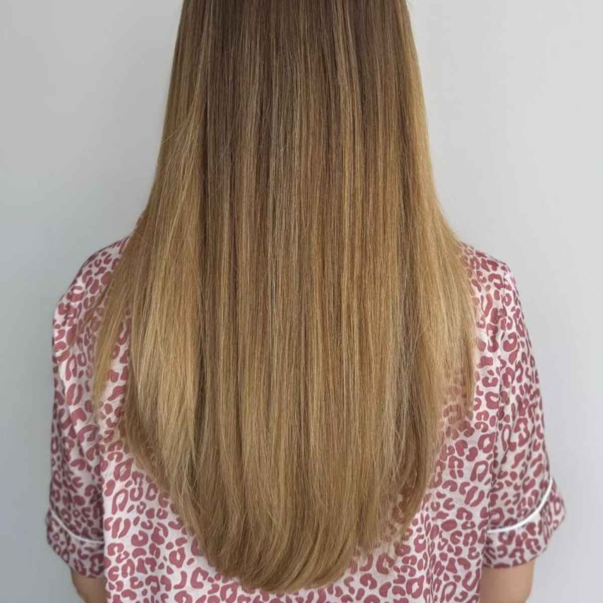 Long, straight, light brown to blonde ombre hair shown from the back of a person wearing a pink and white leopard print top against a plain light gray background.