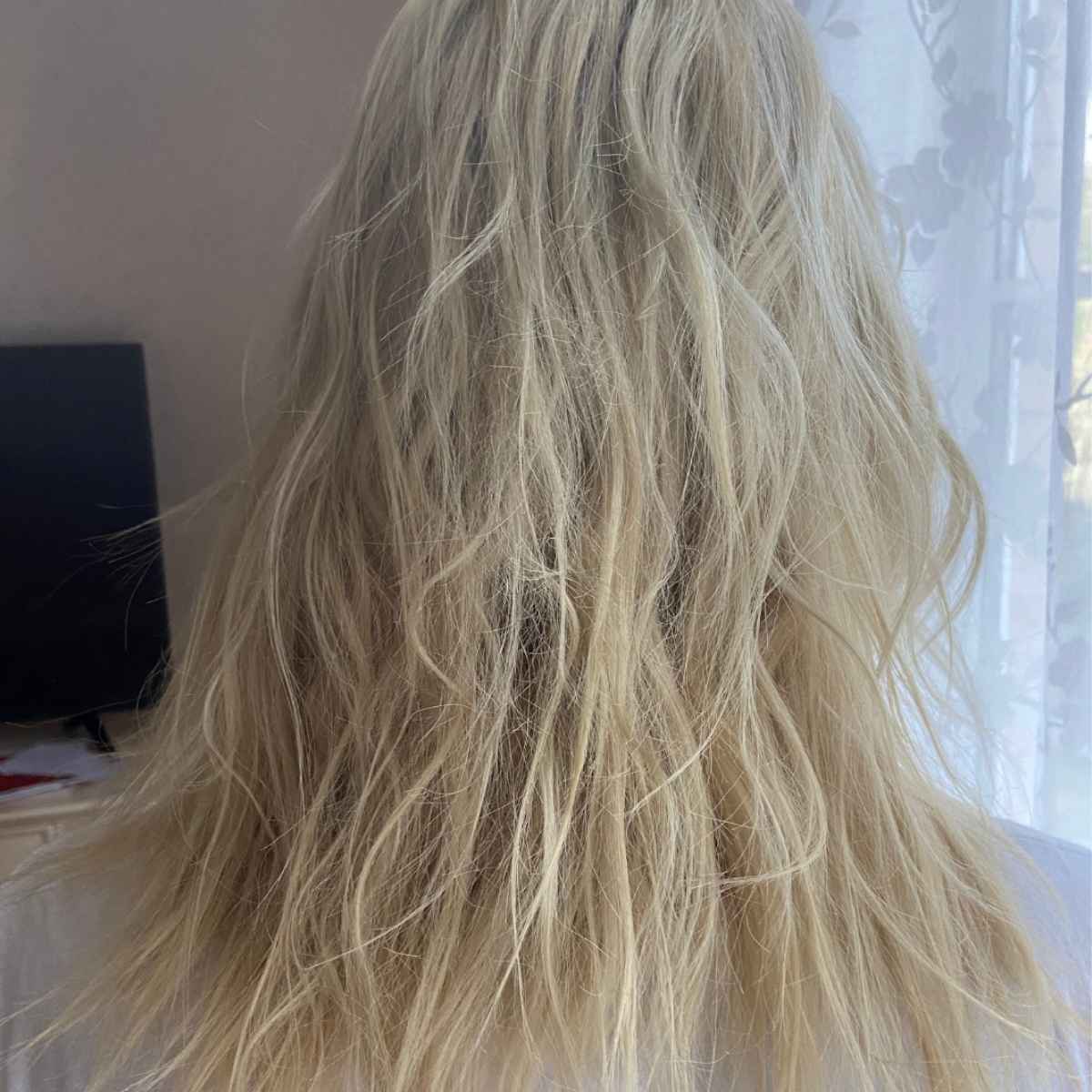 Long, wavy blonde hair shown from the back, filling most of the frame against an indoor background with a curtain and a dark object on the left.